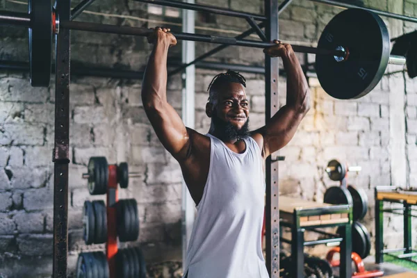 Beyaz gömlekli, güçlü genç Afrikalı erkek sporcu spor salonunda ekipmanla antrenman yaparken ağır halteri kaldırıyor.