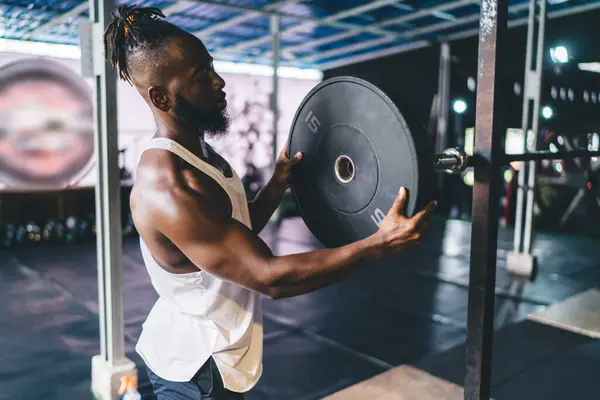 Side view of strong African American bearded male athlete in sportswear lifting and mounting heavy weight plate on metallic bar while working out in gym