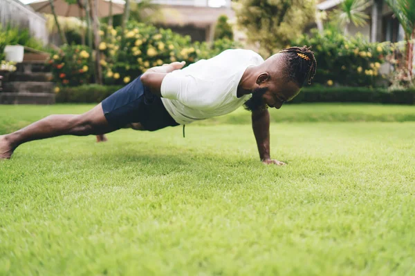 Flexible young African American male doing one arm push up exercise on grassy ground in park while training alone and smiling against blurred green plants