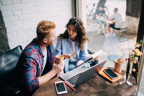 Cheerful young man and woman looking at each other while sitting at table with laptop smartphones and coffee cup in hand while discussing on notes in clipboard during project work in cafe