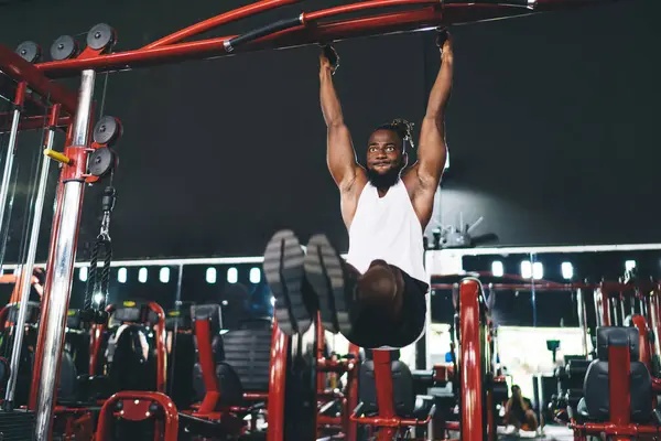 Low angle full body of strong young bearded black male athlete in sportswear doing pull ups on bar with stretched legs and body suspended in modern illuminated gym against dark background