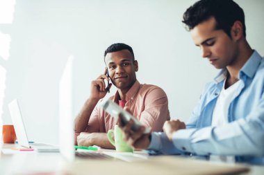 Çok ırklı, entellektüel iş arkadaşları gündelik giysiler içinde masada oturmuş akıllı telefondan konuşuyorlar. Ofisteki kol saatlerini kontrol ediyorlar.
