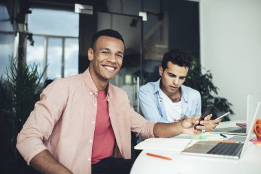 Akıllı telefondan mesaj atan ve kameraya bakan kendinden emin, gülümseyen etnik işçiler. Bu arada endişeli genç meslektaşı not defterindeki bilgilere bakıyor.
