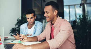 Etnik bir adamın akıllı telefondan mesaj okuması ve ofis masasında genç meslektaşının yanında dizüstü bilgisayarla otururken gülümsemesi.