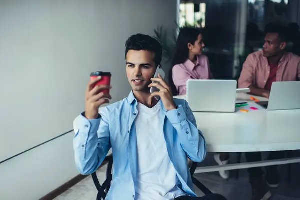 有自信的年轻人坐在办公室 一边用智能手机交谈 一边与同事在模糊的背景下喝咖啡 — 图库照片
