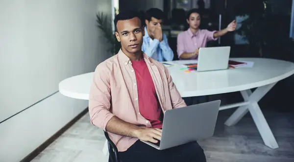不同族裔的办公室工作人员坐在椅子上 手持笔记本电脑 在模糊的背景下看着相机 与积极参加项目讨论的手势相投的同事在一起 — 图库照片
