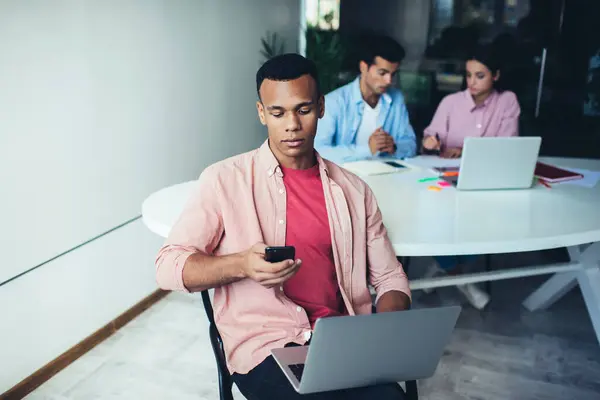 身穿休闲装的年轻非洲裔美国人坐在同事身边 一边在智能手机上浏览社交媒体 一边用笔记本电脑 — 图库照片