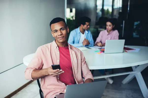 穿着休闲装的积极的非洲裔美国人一边微笑着 一边看着相机 一边在工作场所的同事休息时浏览智能手机和笔记本电脑 — 图库照片