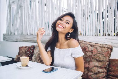 Gülümseyen, omuzları açık genç Asyalı kadın tropikal kafede kahve ve akıllı telefonla masada oturuyor.