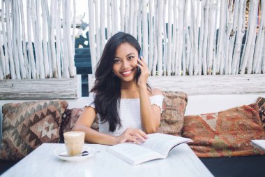 Omuzları açık bluz giyen çekici genç Asyalı kadın elinde kahve fincanıyla masada otururken akıllı telefondan konuşuyor ve tropikal kafede kitap açıyor.