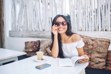 Omzundan çıkmış bluz ve güneş gözlüğü takan neşeli, çekici genç Asyalı kadın tropikal sokak kafesinde elinde kahve ve akıllı telefonla oturmuş kitap okuyor.