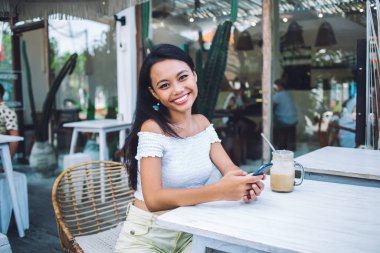 Mutlu, güzel, etnik bir bayan kahve içiyor. Telefonla, terasta oturuyor. Kameraya bakarak gülümsüyor.