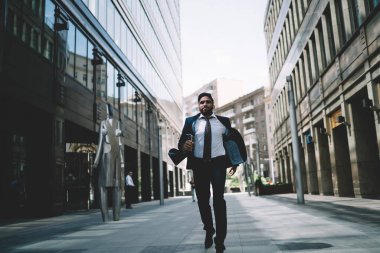 Koyu renk takım elbiseli ve beyaz kravatlı siyah saçlı yönetici erkek şehirde gündüz vakti aceleyle yürüyor.