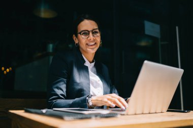 Resmi giyimli ve gözlüklü mutlu bir kadın. Laptop kullanıyor ve kameraya gülümsüyor. Kafe masasında oturuyor ve şehir caddesinde çalışıyor.