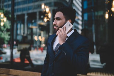 Şık mavi takım elbiseli yakışıklı, düşünceli bir etnik erkek yönetici cep telefonuyla konuşuyor. Arka planda modern binanın cam duvarları var. 