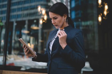 Düşünceli, meşgul bayan girişimci modern şehir caddesinde dijital tabletle iletişim kurarken gözlük takıyor. 