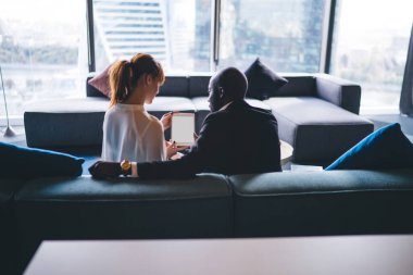 Resmi giyinmiş Afrikalı Amerikalı girişimci ve ofis odasındaki kanepede birlikte oturan ve boş beyaz ekranlı tabletle çalışan genç bayan asistanın arka planı. 