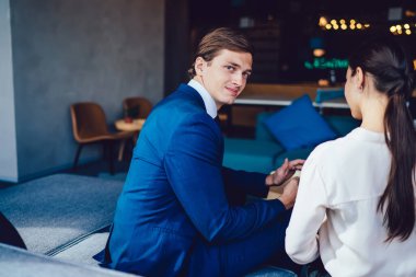 Mavi takım elbiseli zeki erkek yöneticinin çalışma odasındaki kanepede kameraya bakarak pazarlama haberlerini tartışmasının yan görüntüsü.