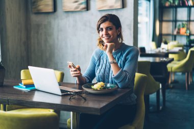 Gündelik kıyafetlerle gezen, akıllı telefonlardan sohbet eden sosyal medyada ara veren, kafede oturmuş kameraya bakan bir kadın.