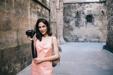 Eski kentteki turizm gezisi sırasında yıpranmış caddede dururken profesyonel fotoğraf makinesiyle gülümseyen sıska genç bir kadın.