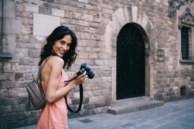 Modern fotoğraf makineli kadın turistin yan görüntüsü neşeyle gülümserken antik kentteki taş binanın yanında duruyor.