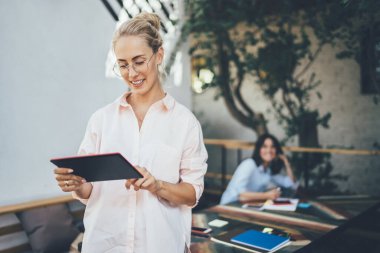 Gözlüklü neşeli bayan, gülümsüyor ve tablet kullanıyor. Masanın yanında duruyor ve kahve terasında meslektaşını bulanıklaştırıyor.