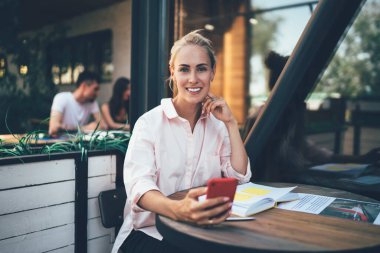 Çekici, gülümseyen, serbest çalışan bir kadın. Kafe terasında küçük bir masada oturuyor ve yeni bir proje için bilgi ararken cep telefonunu kullanıyor.