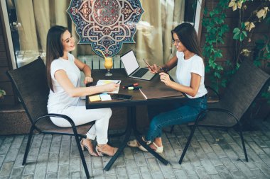 Neşeli, neşeli, zeki kadınların yeni başlatılan proje ile ilgili anlaşmaya varmak için işbirliği yapmalarına ve açık kafede otururken pazarlık için cihazlar kullanmalarına yandan bakın.