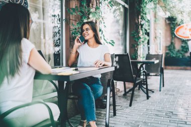 Başarılı bir serbest çalışan, gözlüklü, büyüleyici bir şekilde gülümseyen, telefon görüşmesi yapan bir kadın, açık hava kafede uzaktan bakan bir bayan meslektaşıyla oturuyor.