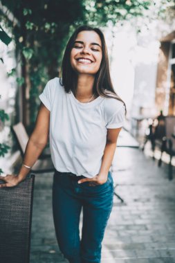 Gündelik giysiler içinde gülümseyen güzel bir kadın. Kot pantolon cebinde. Kafe terasında dikilip yaz günü kameraya gülüyor.