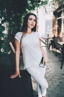 Beyaz elbiseli, kendine güveni tam bir genç bayan. Elinde telefon, güneşli bir günde açık hava barında dinlenirken masaya yaslanıyor.