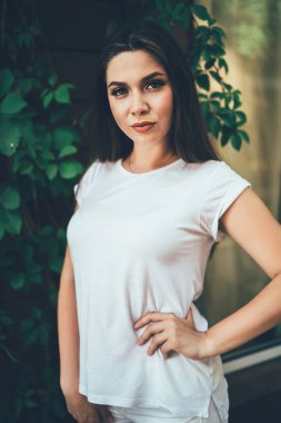 Koyu renk saçlı, kolları çıplak, bembeyaz giyinmiş, ağaç gövdesinin karşısında duran yeşil yapraklı bir kadın.