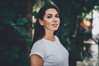 Kendinden emin, koyu renk saçlı, dolgun dudaklı, beyaz gömlekli, arka planda yeşillik olan bir kadın.