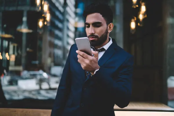 Serious adult Hispanic executive man in elegant clothes focusing on smartphone screen and thinking while standing against window wall in contemporary office building