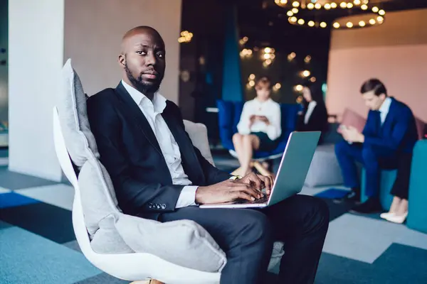 Side view of pensive smart serious black manager in corporate outfit working on laptop while sitting alone on armchair separately from colleagues looking away