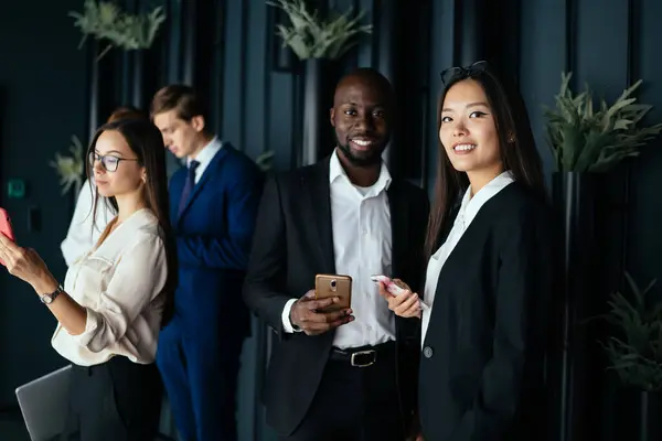 Low angle of adult diverse entrepreneurs in elegant clothes smiling at camera while standing beside colleagues against black striped wall with potted green plants and interacting with smartphones in conference hall during break in work