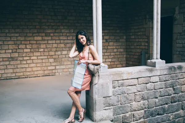 Young woman in peach dress leaning on column near staircase looking at camera while having sightseeing tour with map in old city