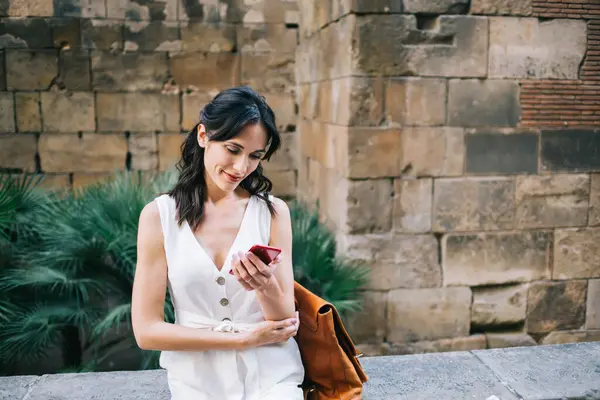 Siyah saçlı, kısa beyaz elbiseli, büyük çantalı bir kadın. Eski binanın karşısındaki kırmızı cep telefonundan mesaj yolluyor.