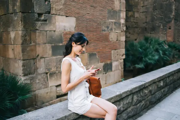 Siyah saçlı, beyaz kolsuz giysili neşeli bayan yazın taş korkulukta oturan cep telefonunu izliyor.