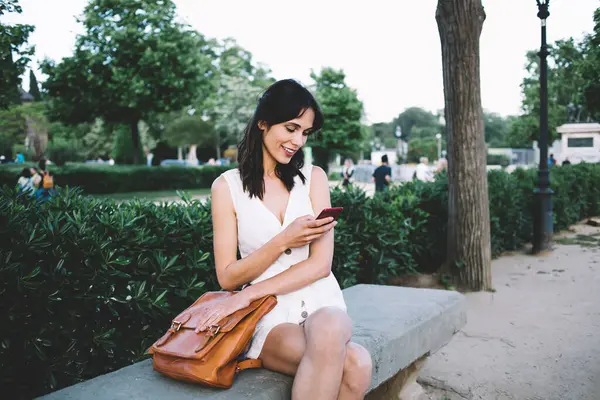 Beyaz elbiseli neşeli esmer kadın ve şık çantalı cep telefonu yeşil çitli bankta oturuyor. 