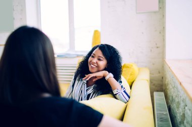 Genç ve çekici Afro-Amerikan bayan rahat bir şekilde sarı kanepeye uzanmış, eline yaslanmış ve dikkatle uzun saçlı arkadaşını dinliyor. 