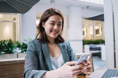 Kafede, tahta masada beyaz ekranlı kalem ve dizüstü bilgisayarla otururken akıllı telefondan mesaj atan mutlu Asyalı genç serbest yazar.