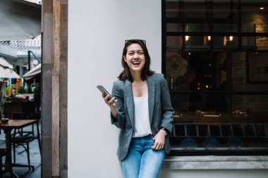 Güzel, güzel, mutlu, resmi giyinmiş, akıllı telefon kullanan, şehrin caddesindeki kameraya gülen bir kadın.