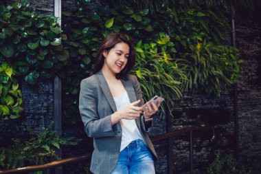 Mutlu, genç, gülümseyen, şık kıyafetler içinde güzel bir Asyalı kadın. Parkta, gri ceket ve kot pantolonla telefonla mesajlaşıyor. 