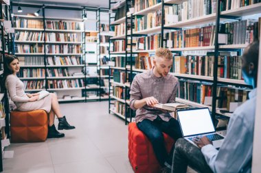 Kitapları olan, gülümseyen ve çok ırklı sınıf arkadaşlarına bakan, boş ekranlı dizüstü bilgisayar kullanan, kitaplıkların yanında oturan ve kütüphanede ödev yapan genç bir kadın.