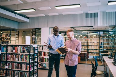 Genç, neşeli, çok kültürlü erkek öğrenciler gündelik kıyafetlerle edebiyatı tartışırken ayakta durup kitapları tutarken ve kütüphanede neşeyle konuşurken. 
