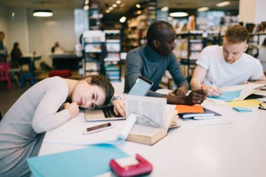 Günlük giysiler içinde yorgun bir kadın dizüstü bilgisayarda uyuyor iş arkadaşlarının yanında ofis binasında projeyi tartışan defterlerle.