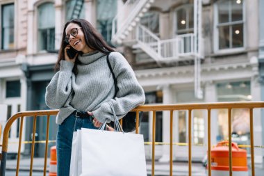 Kazak giyen, bariyere yaslanmış, alışveriş torbalarıyla konuşan ve New York 'ta cep telefonuyla konuşan neşeli, genç bir kadın.