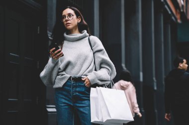 New York 'un caddesinde dikilirken şık tatlı giyinmiş, gözlüklü, alışveriş çantalı ve cep telefonlu güzel bir kadın.
