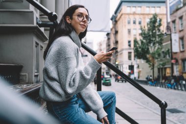 Gri kazaklı ve gözlüklü yetişkin bir kadının sokak merdivenlerinde oturmuş elinde akıllı bir telefonla gülümsemesi. 